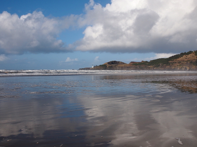 [Puffy white clouds in a blue sky are reflected on the water lying just above the surface of the sand. A strip of land coming from the right is also reflected in the water.]